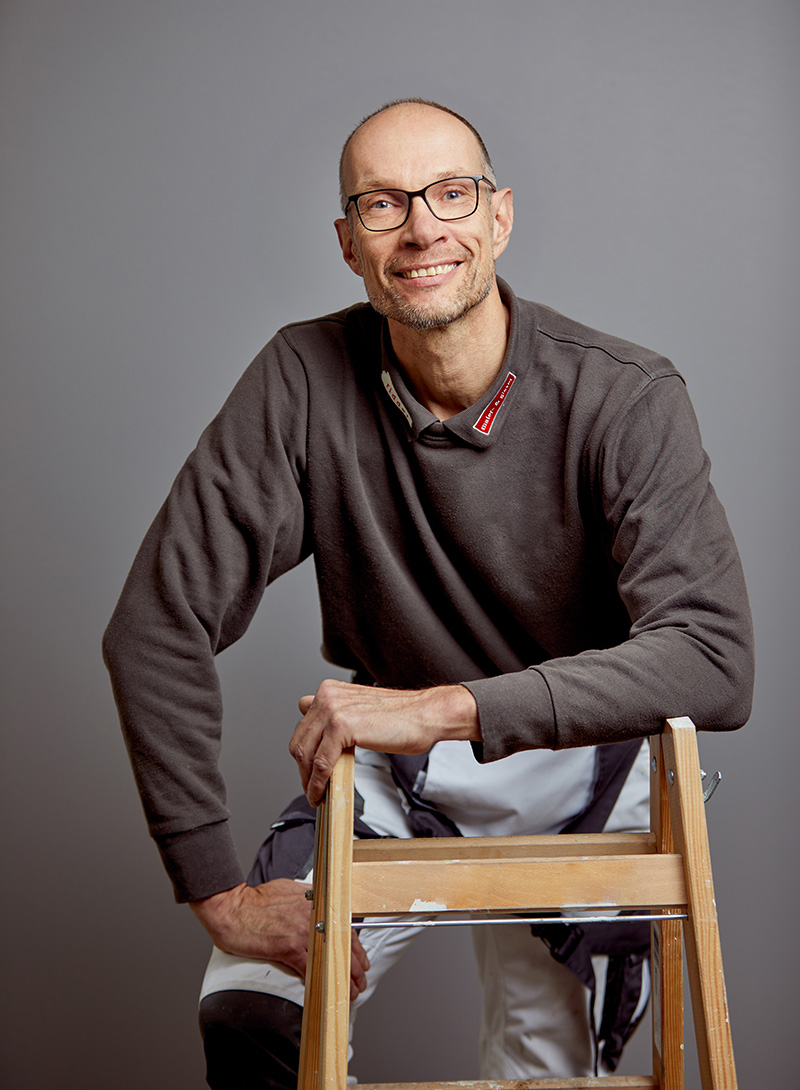 Mann im Maleroutfit lehnt an der Leiter, Brille und lächelt in die Kamera