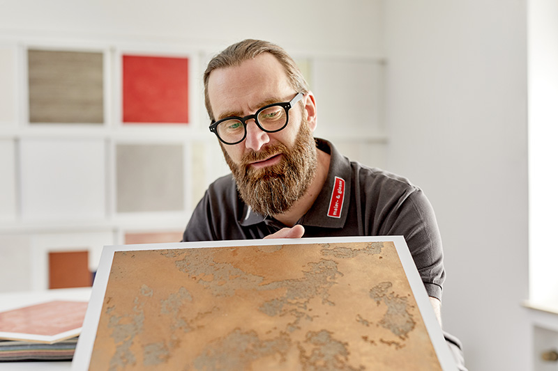 Mann zeigt eine Farbplatte in die Kamera, er schaut es sich an. Trägt Brille und Bart