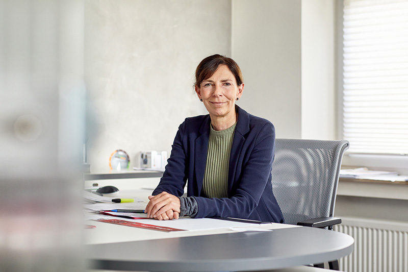 Frau im blauen Jacket sitzt am Schreibtisch und lächelt in die Kamera