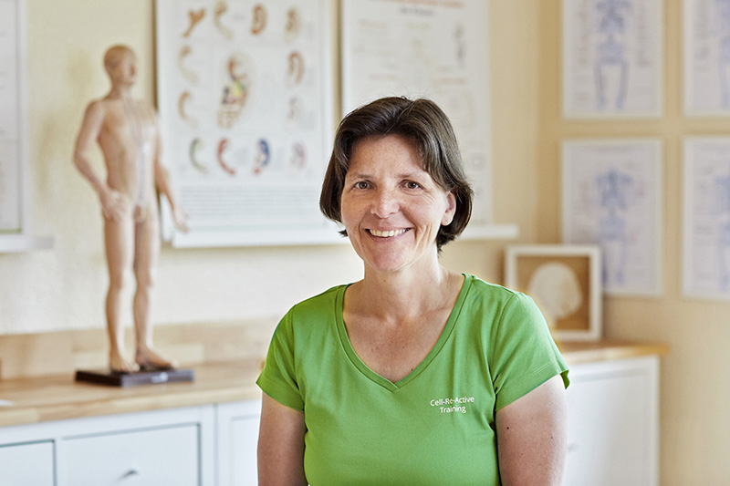 Frau im grünen Shirt mit der Aufschrift "Cell-Reactive Training" lächelt in die Kamera. Im Hintergund erkennt man unscharf eine nackte Figur. Es isr eine Heilpraktiker-Praxis