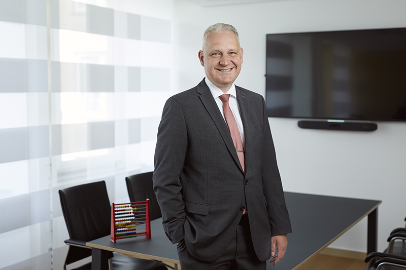 Mann im Anzug steht vor Bürotisch, eine Hand in der Tasch, lächelt