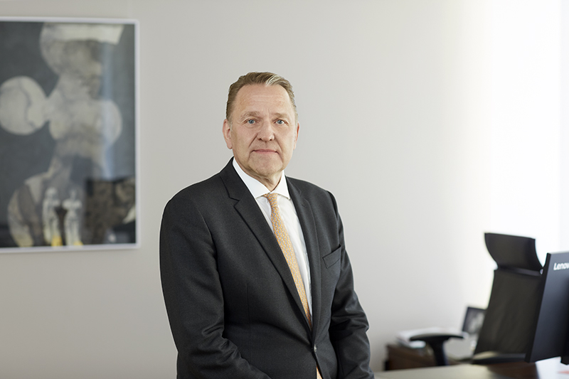 Mann im Anzug sitz auf Schreibtisch, weißer Hintergrund mit unscharfem Bild links am Bildrand