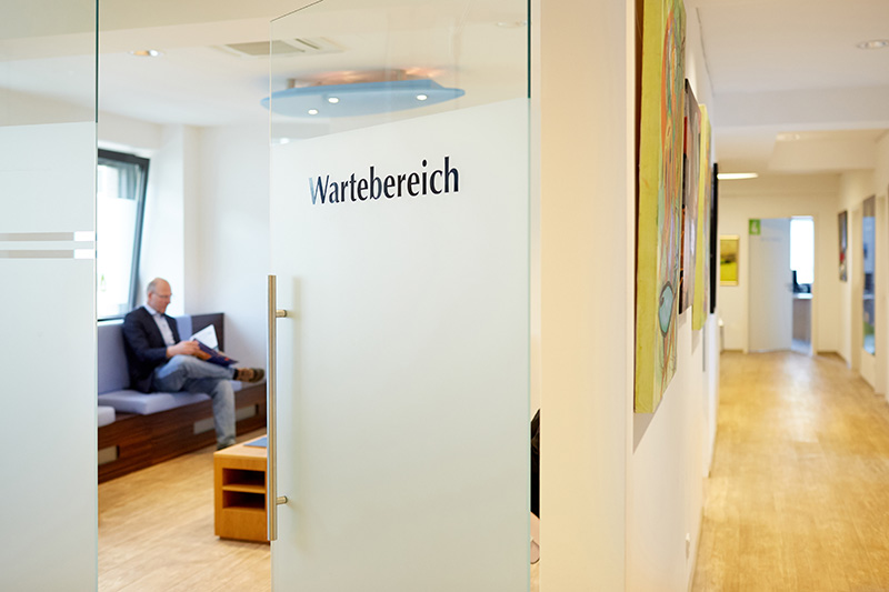 Hinter einer Tür mit der AUfschrift "Warteberich" sitzt ein Mann lesend im Wartzimmer einer Arztpraxis